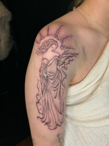 Tattoo of an angel with flowing robes and a halo on a shoulder.