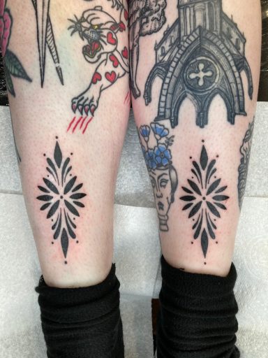 Black ink tattoos featuring intricate symmetrical designs on each calf.